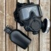 Black gas mask with attached filter and a black canteen on wooden surface.